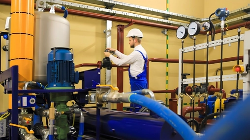 Worker in a hard hat among blue, orange, and red industrial pipes and pressure gauges