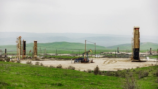 Multiple pump jacks spread across a green oilfield on a cloudy overcast day with low hills in the distance