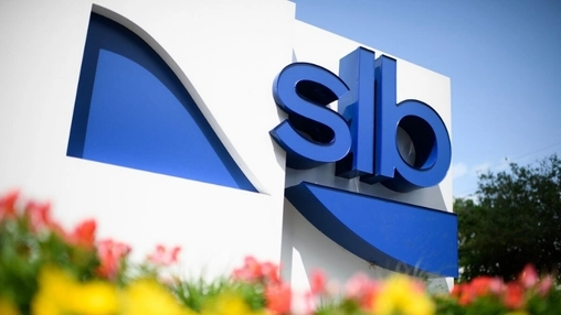 Close-up of SLB blue branded sign on white wall with red and yellow flowers in foreground and trees behind