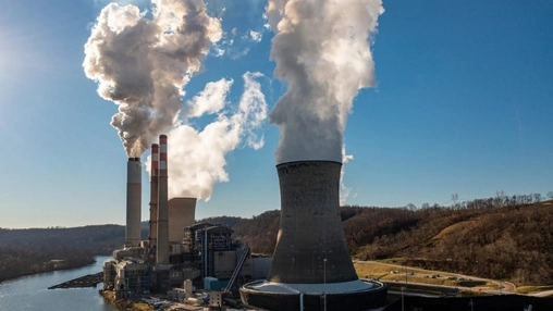 Industrial power facility with active cooling tower and twin smokestacks emitting emissions beside a river