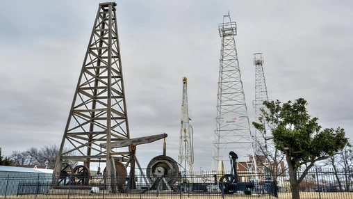 Wooden oil derrick and steel drilling towers at an outdoor oil and gas heritage site under an overcast sky