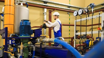 Worker in a hard hat among blue, orange, and red industrial pipes and pressure gauges