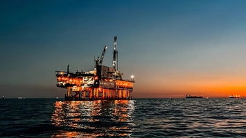 An illuminated offshore oil platform at dusk reflected on calm open water with vessels in the background