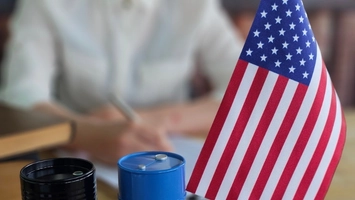 US flag on a desk next to small black and blue oil drum models, with a person working in the background